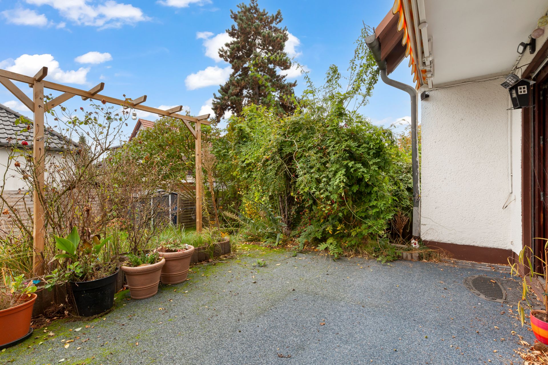 Terrasse mit Blumentöpfen, Holzgitter und viel Grün neben einem Wohnhaus.