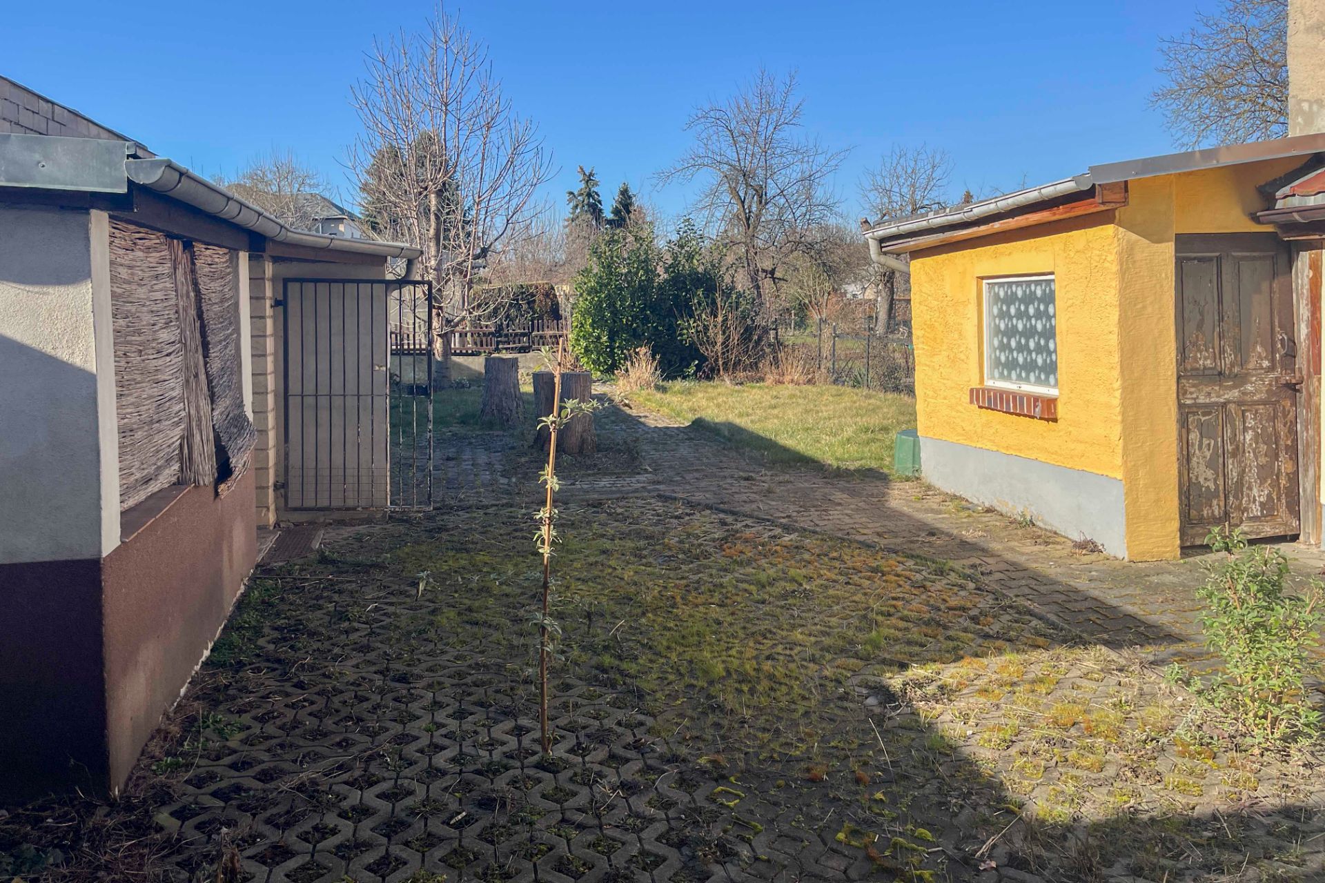 Gepflasterter Hof mit gelbem Anbau, Rasenfläche und Bäumen im Hintergrund.