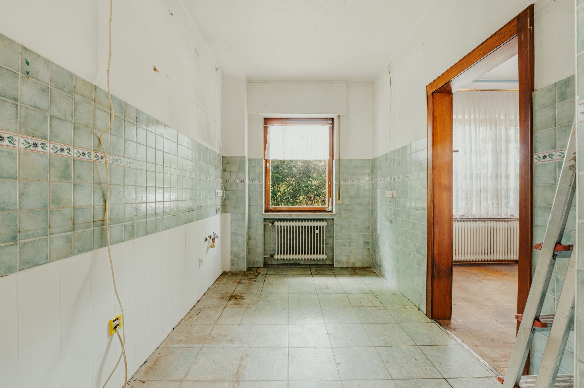 Leerer Raum mit grünen Fliesen, Fenster, Heizkörper und Leiter im Vordergrund.