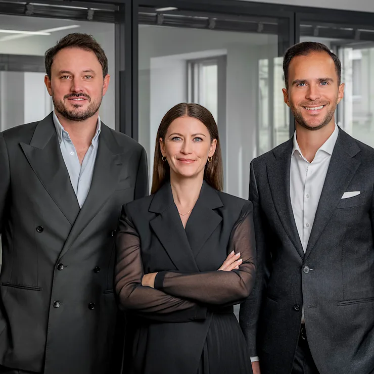 Drei Personen in Businesskleidung stehen in einem modernen Büro mit Glaswänden.