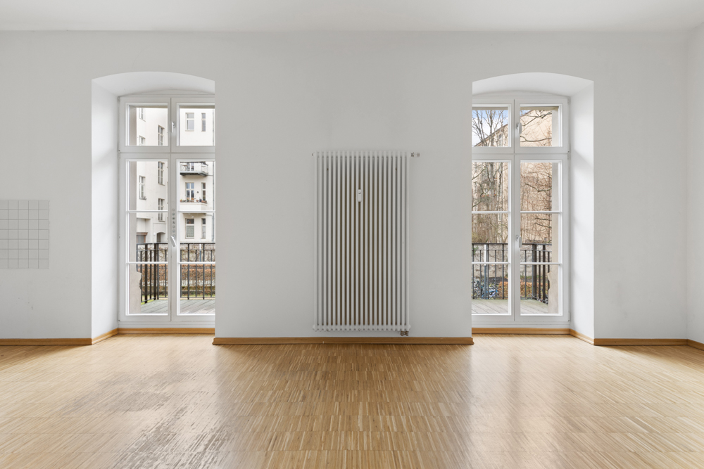 Heller Raum mit Parkettboden, zwei großen Fenstern und einem Heizkörper an der Wand.