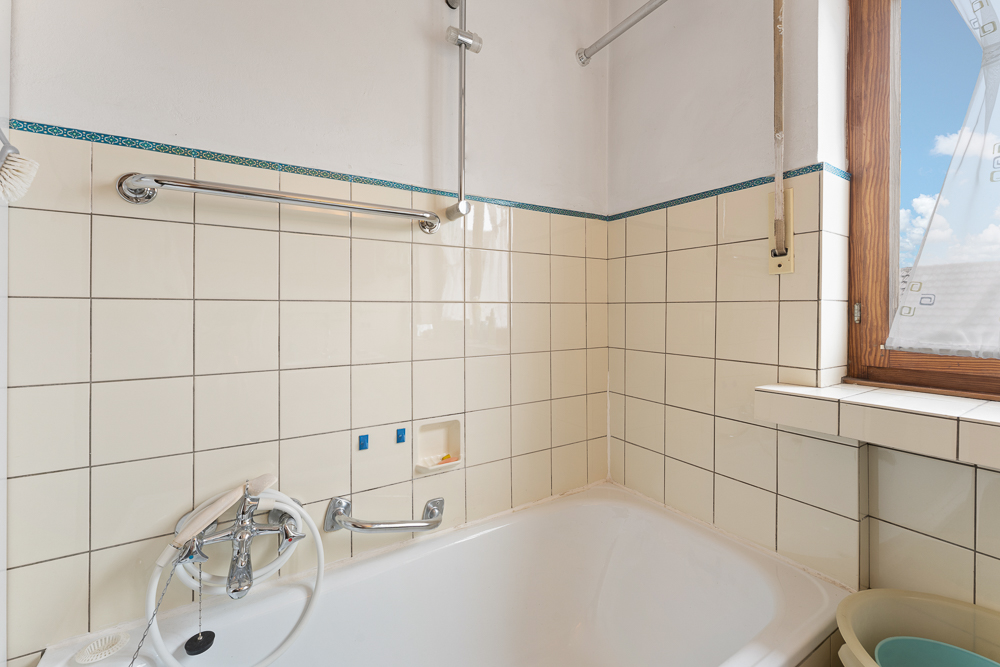 Ecke eines Badezimmers mit Badewanne, beigen Fliesen, Fenster und Handtuchhalter.