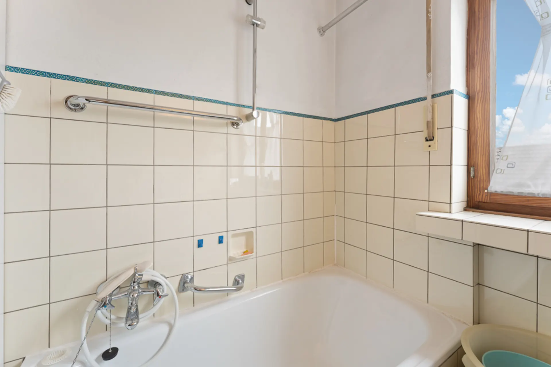 Ecke eines Badezimmers mit Badewanne, beigen Fliesen, Fenster und Handtuchhalter.