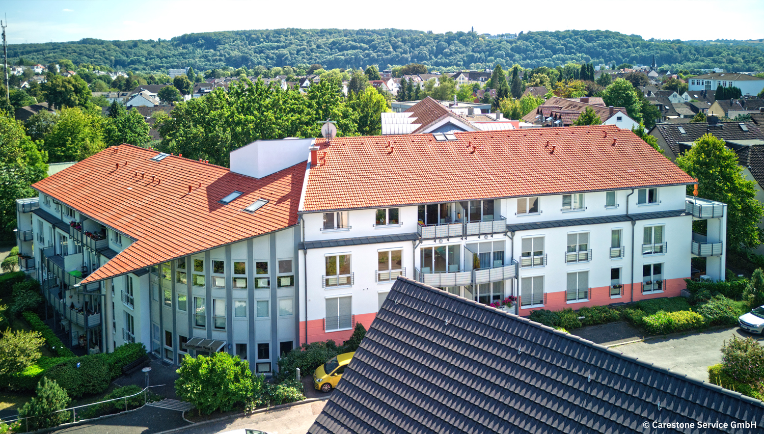 Wohngebäude mit rotem Dach, Balkonen und umgeben von Bäumen, aus der Vogelperspektive.