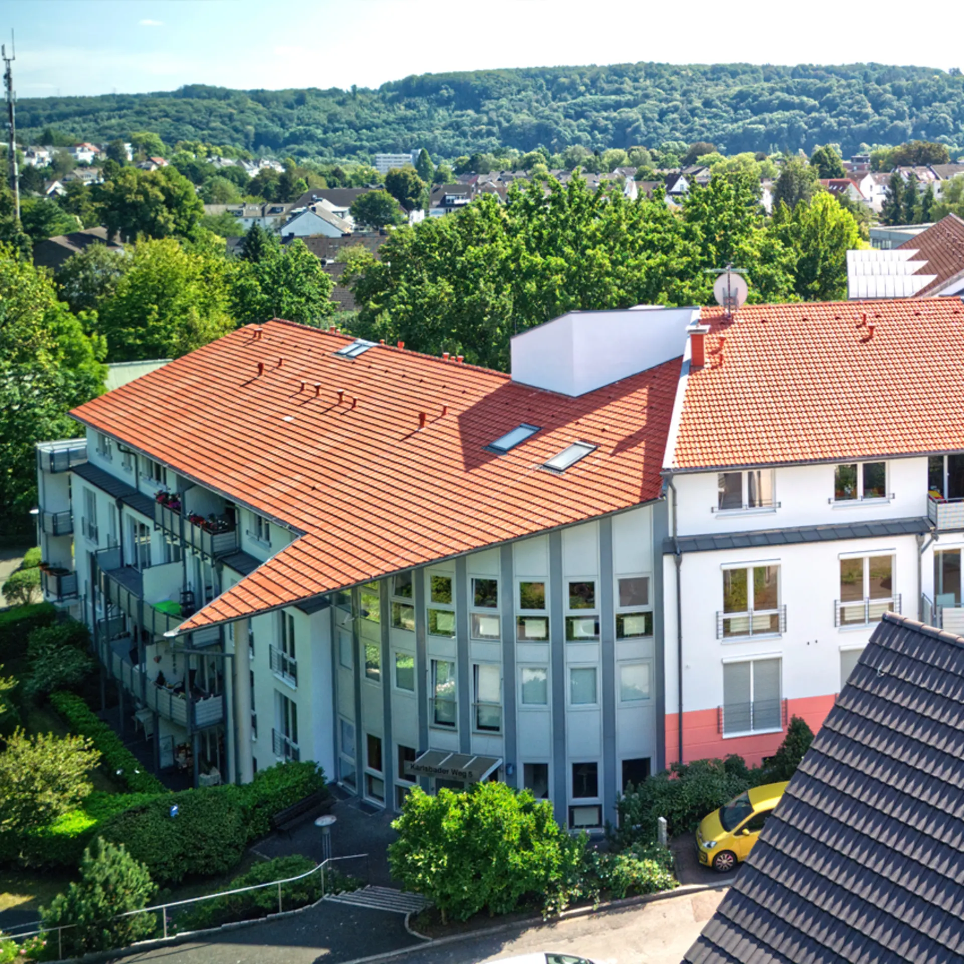 Wohngebäude mit rotem Dach, Balkonen und umgeben von Bäumen, aus der Vogelperspektive.