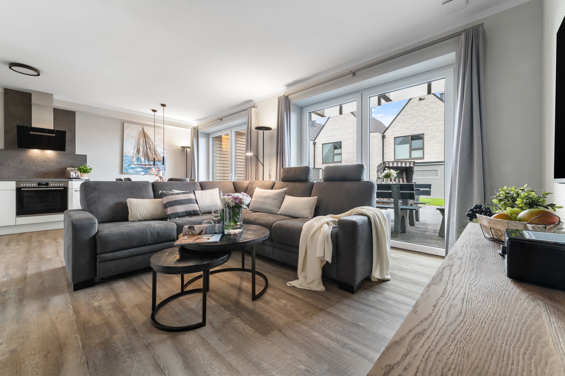 Modernes Wohnzimmer mit grauem Sofa, Couchtisch, Blick auf Küche und große Fenster mit Terrasse.