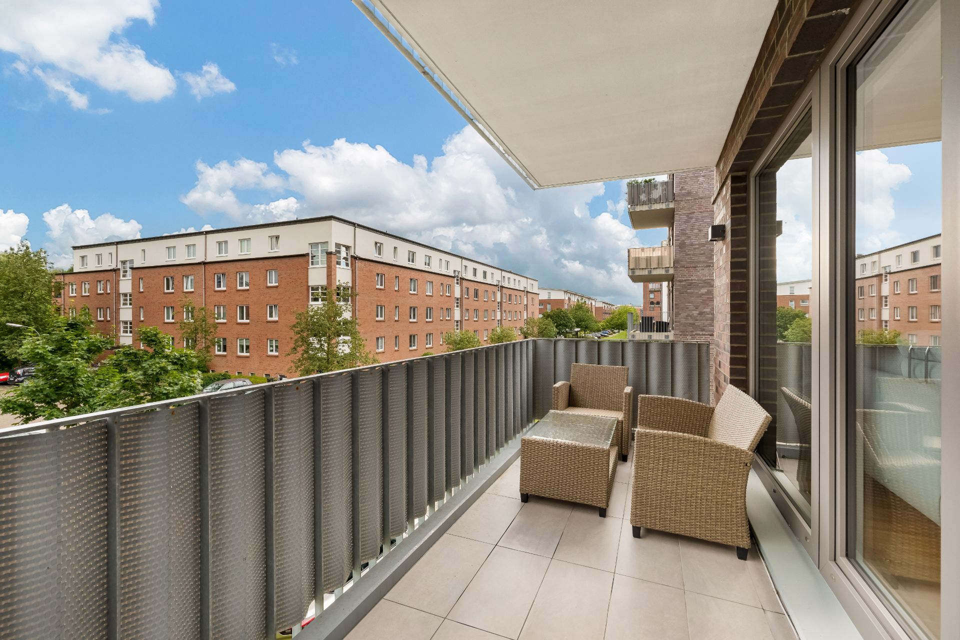 Balkon mit zwei Korbstühlen, Tisch und Blick auf Wohngebäude an einem sonnigen Tag.