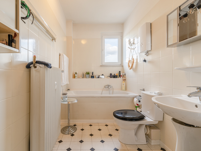 Helles Badezimmer mit Badewanne, Toilette, Waschbecken und Fenster, gefliest, mit einigen Hygieneartikeln.