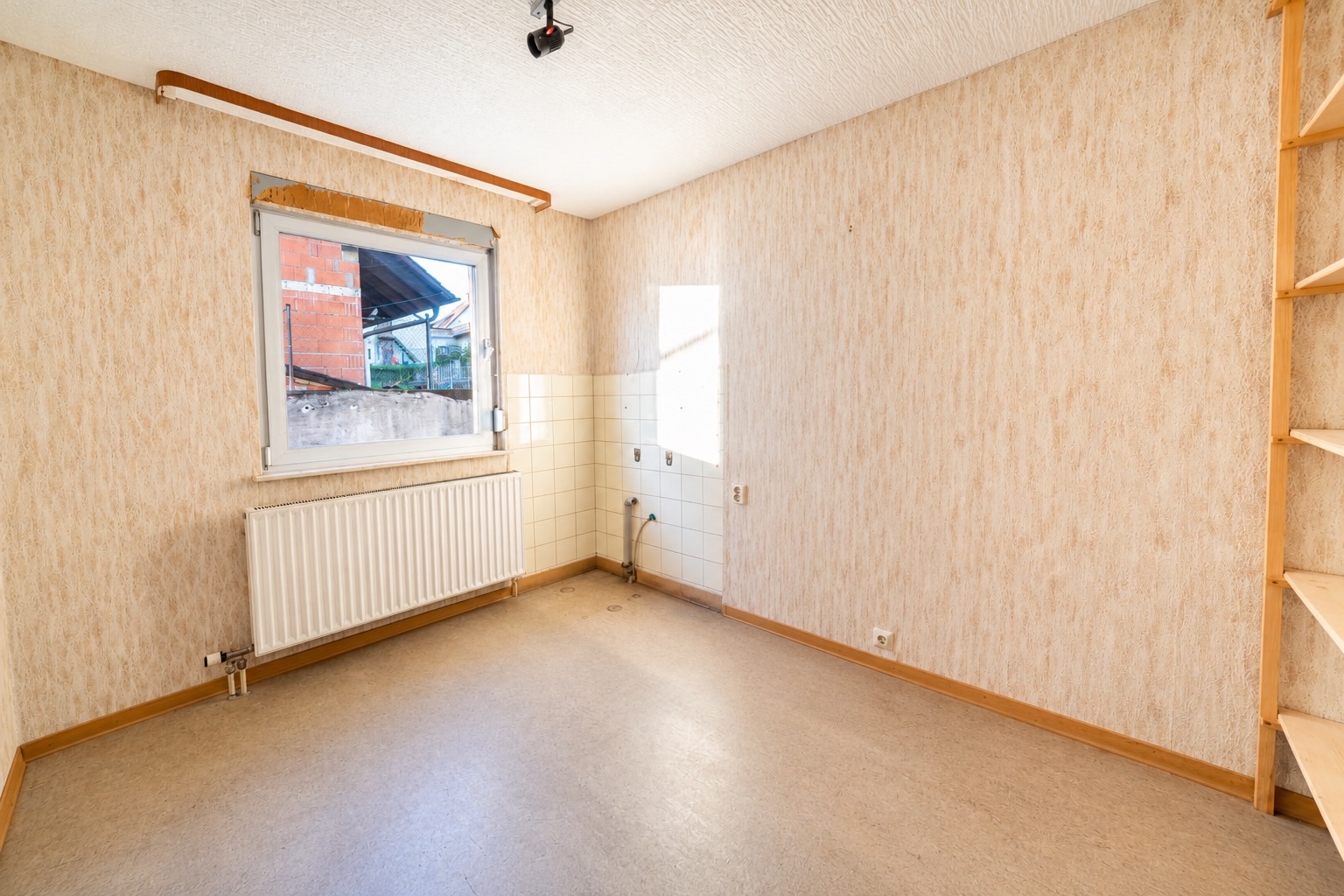 Leerer Raum mit Fenster, Heizkörper, hellem Boden und Regalen an der rechten Wand.