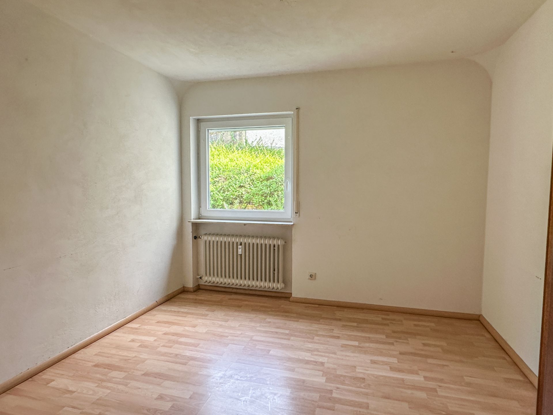 Leeres Zimmer mit hellem Laminatboden, weißer Wand, Fenster und Heizkörper.