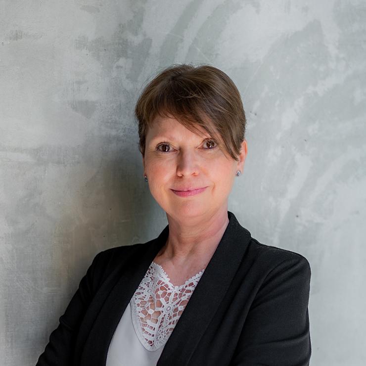 Frau mit kurzem Haar und schwarzem Blazer steht vor grauer Wand.