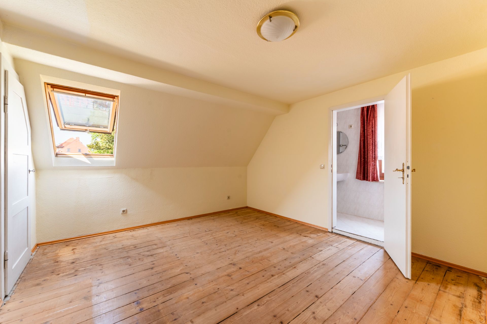 Helles, leeres Zimmer mit Holzboden, Dachfenster und offenem Zugang zu einem Badezimmer.
