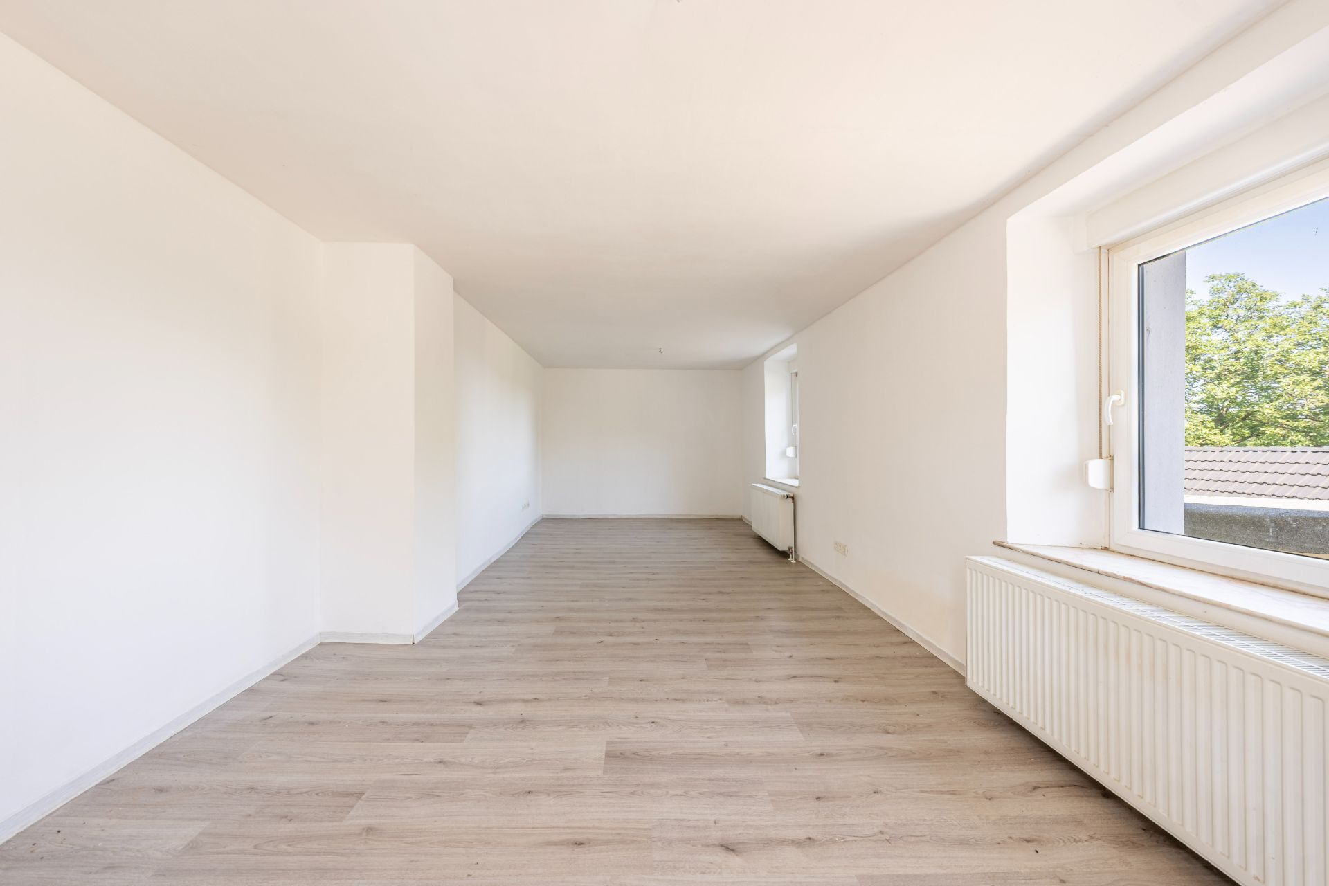 Helles, leeres Zimmer mit großem Fenster, weiß gestrichenen Wänden und hellem Holzboden.