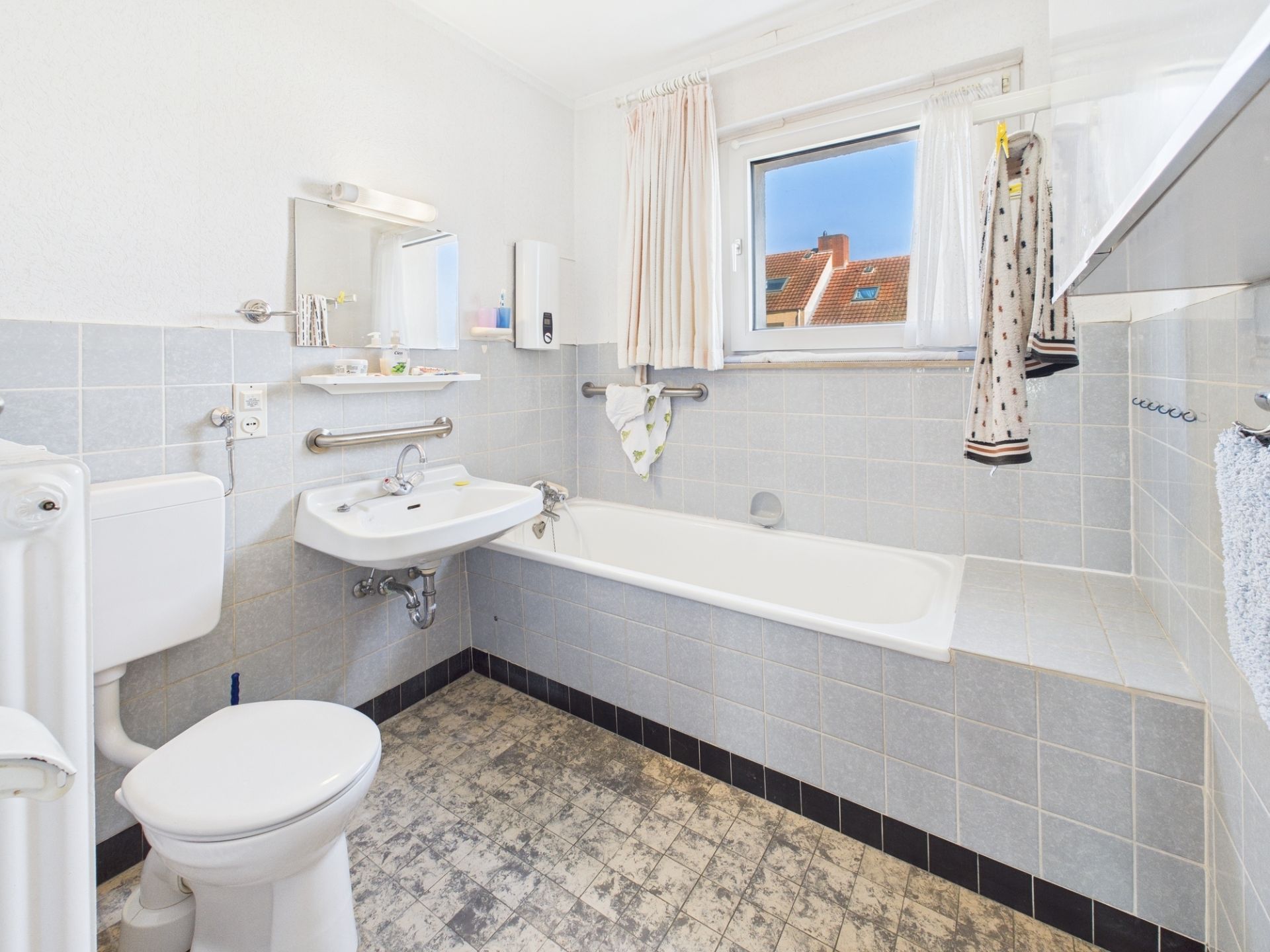 Helles Badezimmer mit Badewanne, Waschbecken und WC, Fenster mit Blick auf Dächer.