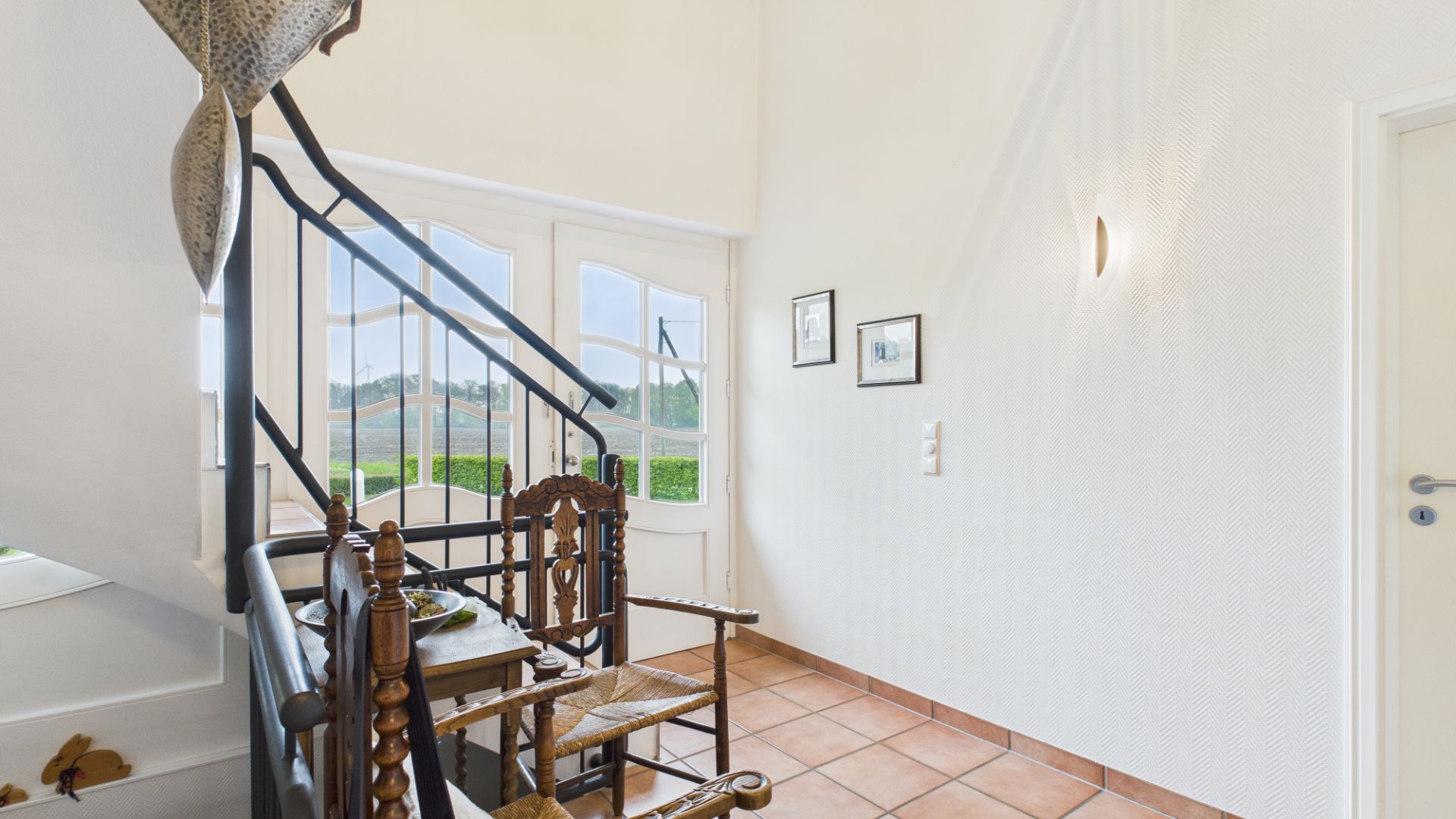 Heller Flur mit Wendeltreppe, Holztisch und zwei Stühlen, Fliesenboden und Wandbildern.