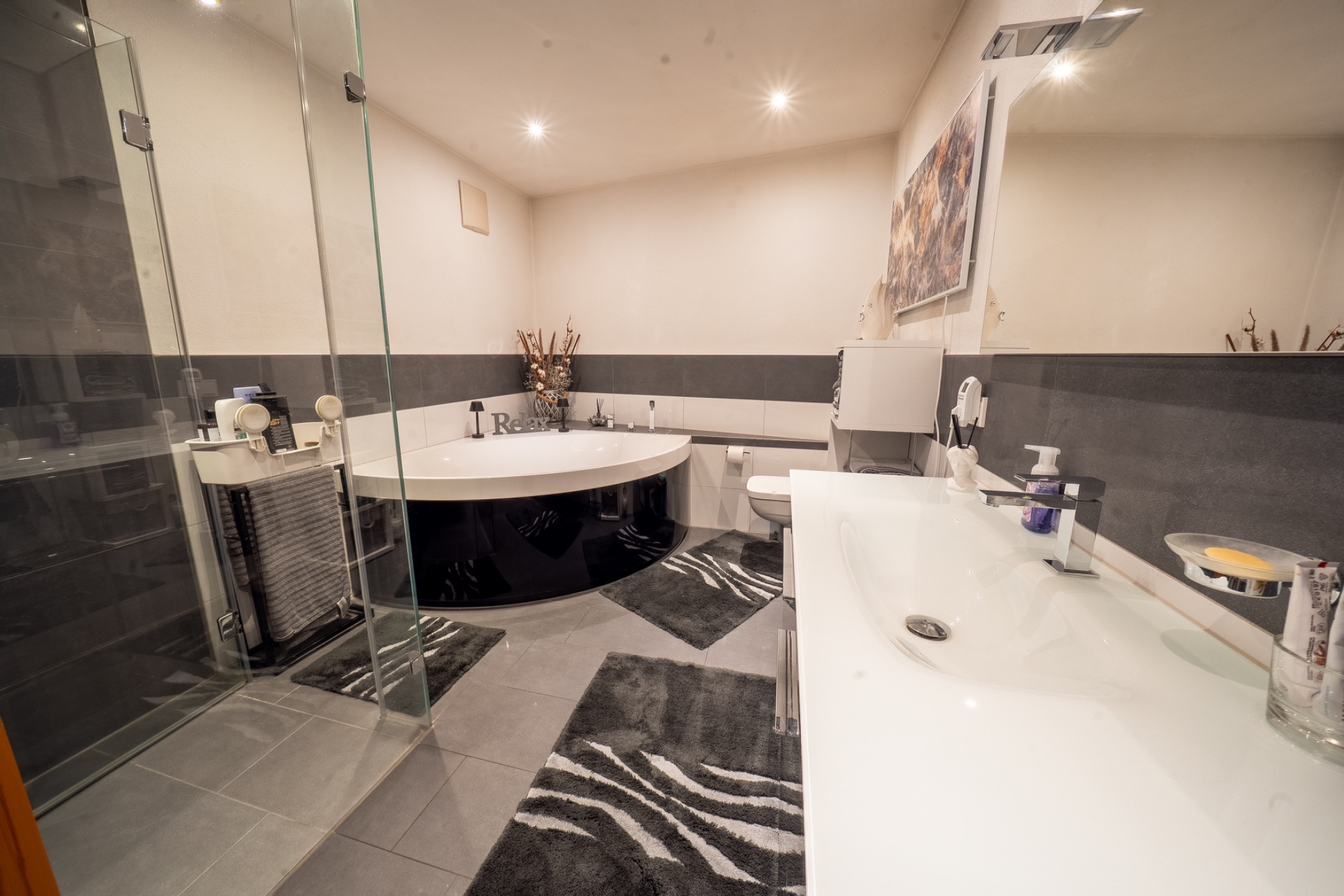 Modernes Badezimmer mit runder Badewanne, Dusche, langem Waschbecken und grauen Teppichen.