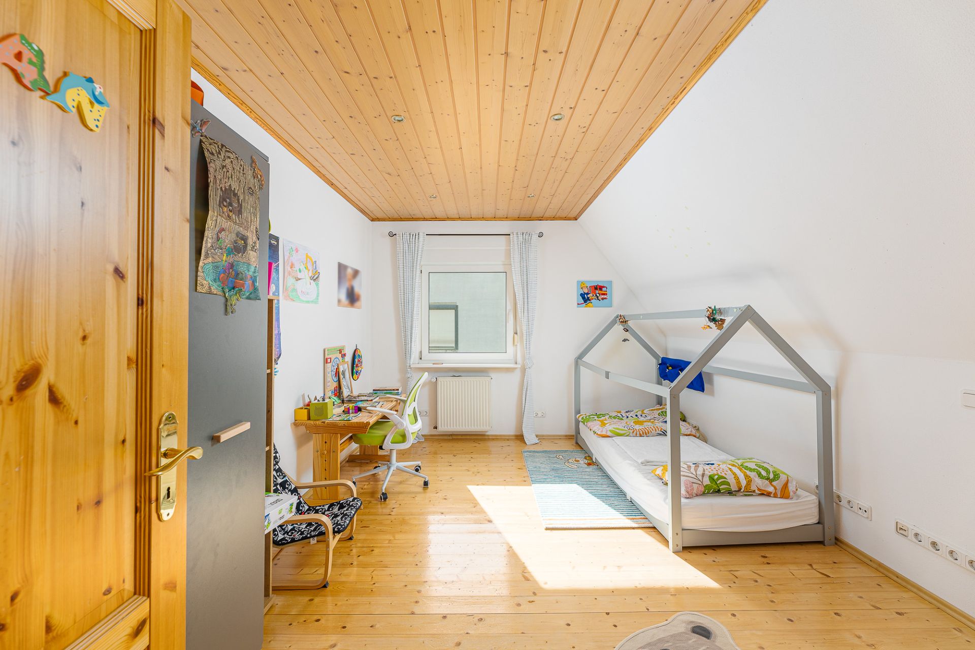 Helles Kinderzimmer mit Holzboden, Holzbett und Schreibtisch unter einem Fenster.
