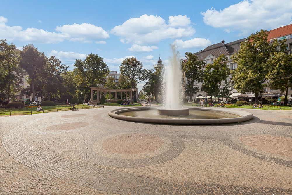 Großer Springbrunnen auf einem gepflasterten Platz, umgeben von Bäumen und Gebäuden.