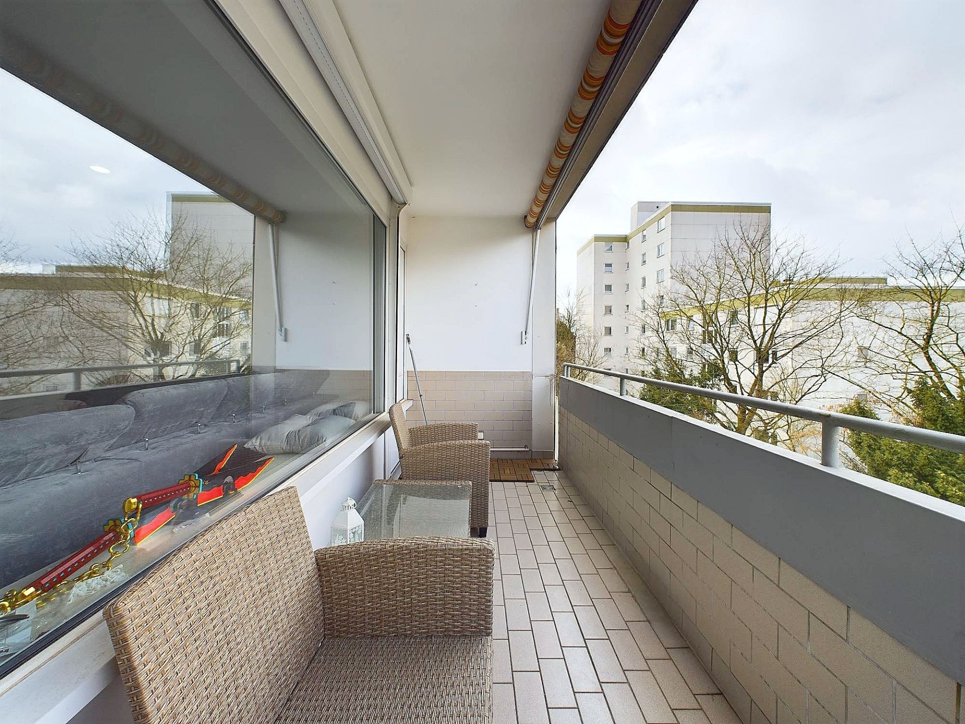 Balkon mit zwei Korbstühlen, Glastisch und Ausblick auf benachbarte Wohngebäude.