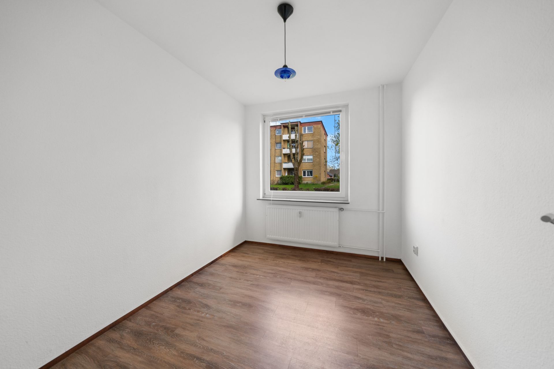 Kleines, leeres Zimmer mit weißem Wänden, Holzfußboden und Fenster mit Blick auf ein Mehrfamilienhaus.