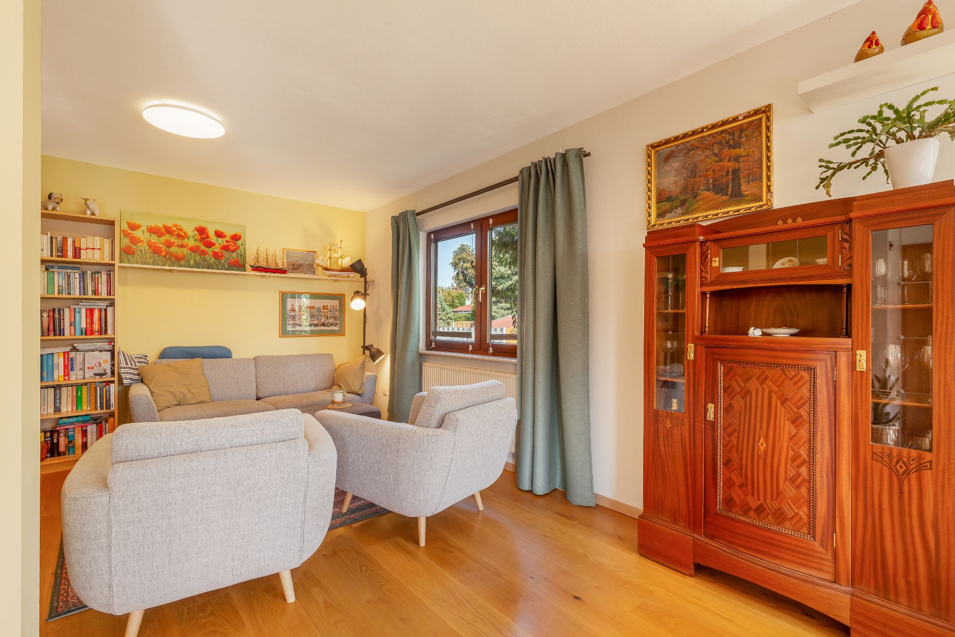Helles Wohnzimmer mit grauem Sofa, Sessel, Bücherregal und Holzbuffet, Fenster mit Vorhängen.