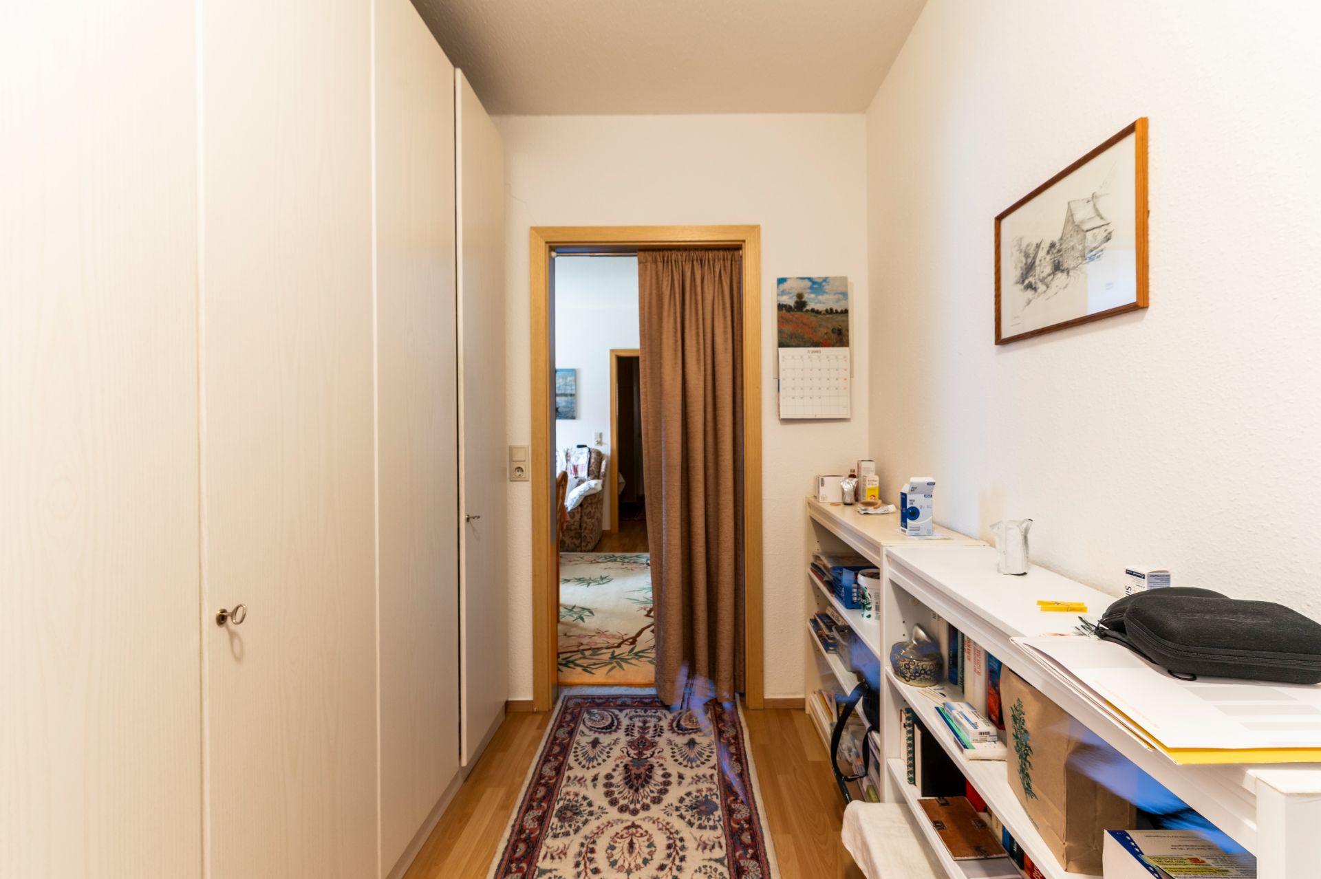 Schmaler Flur mit Teppich, Einbauschrank links und Regal rechts, Blick in ein weiteres Zimmer.