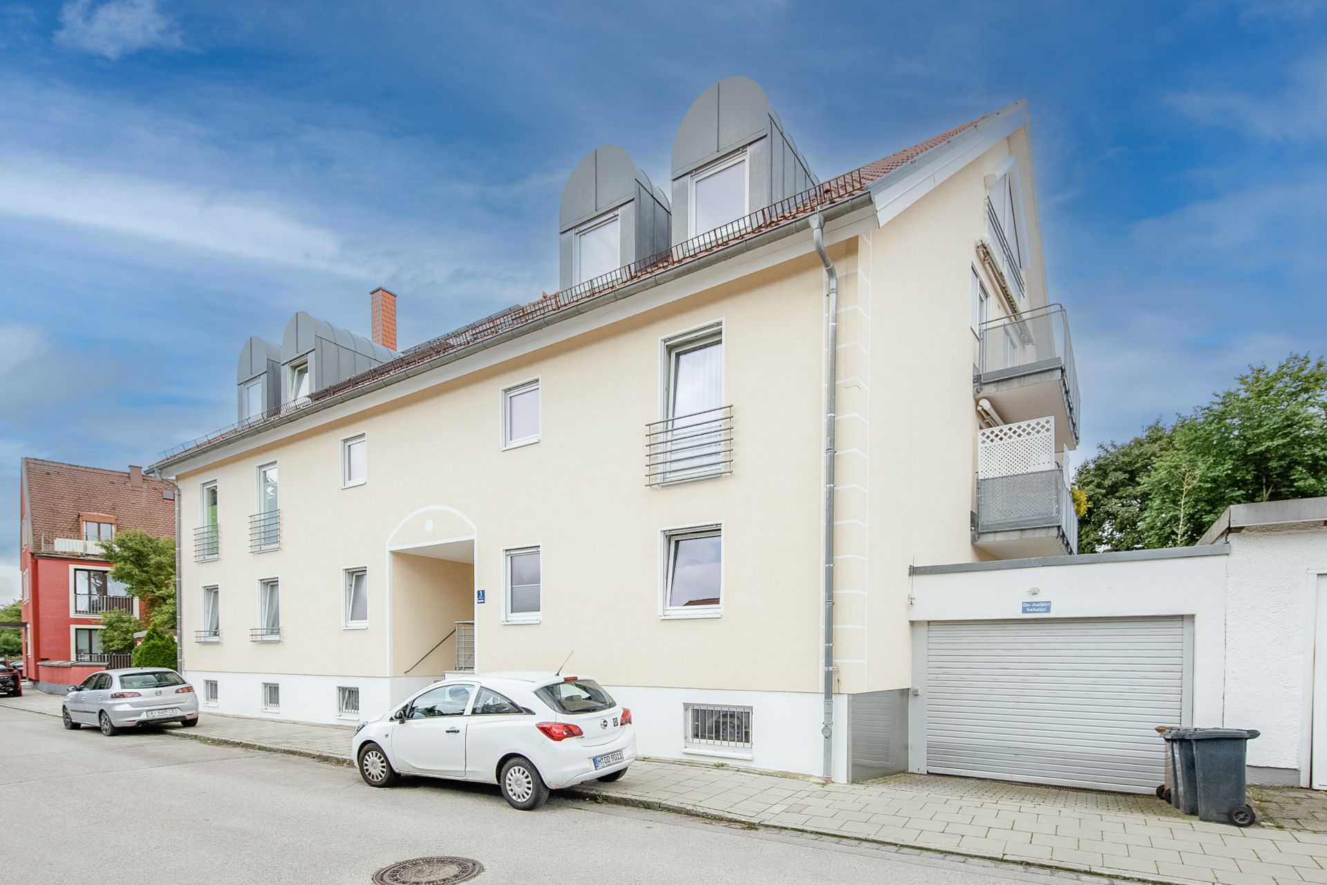 Helles Wohnhaus mit mehreren Fenstern, Dachgauben und Garage an einer ruhigen Straße.