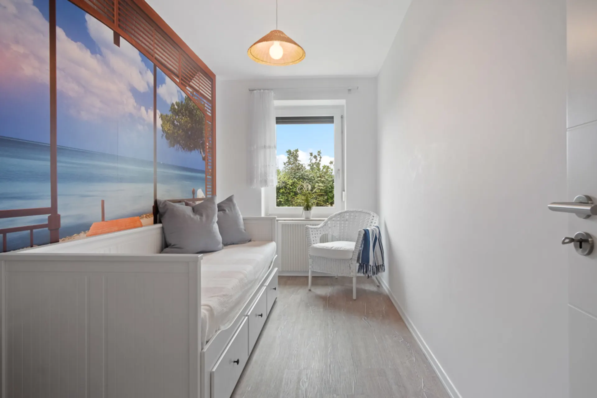 Helles Zimmer mit Tagesbett, Wandbild von Meerblick, weißem Stuhl und Fenster mit Ausblick auf Bäume.