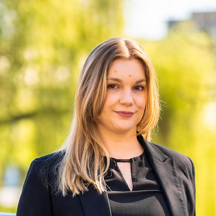 Eine Frau mit langen, blonden Haaren steht draußen vor grünem, unscharfem Hintergrund.