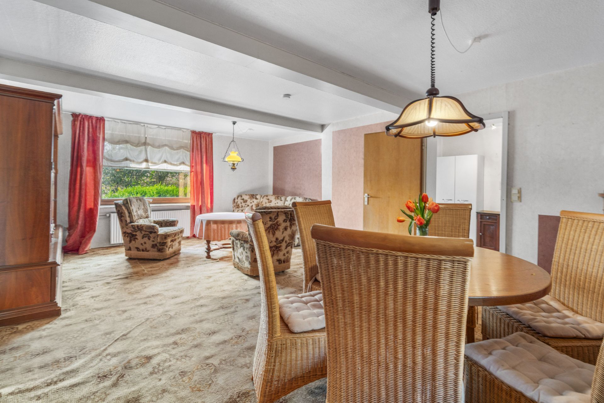 Wohnzimmer mit Esstisch, Stühlen, Sofa, Sessel, Holzmöbeln und großen Fenstern mit roten Vorhängen.