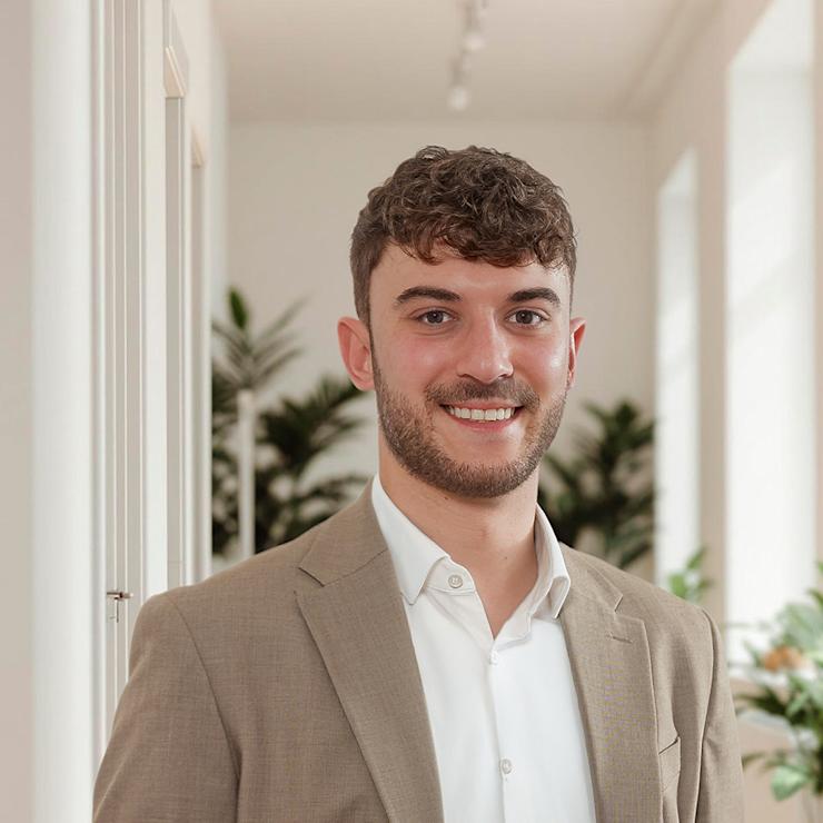 Ein Mann im beigen Anzug steht in einem hellen Raum mit Pflanzen im Hintergrund.