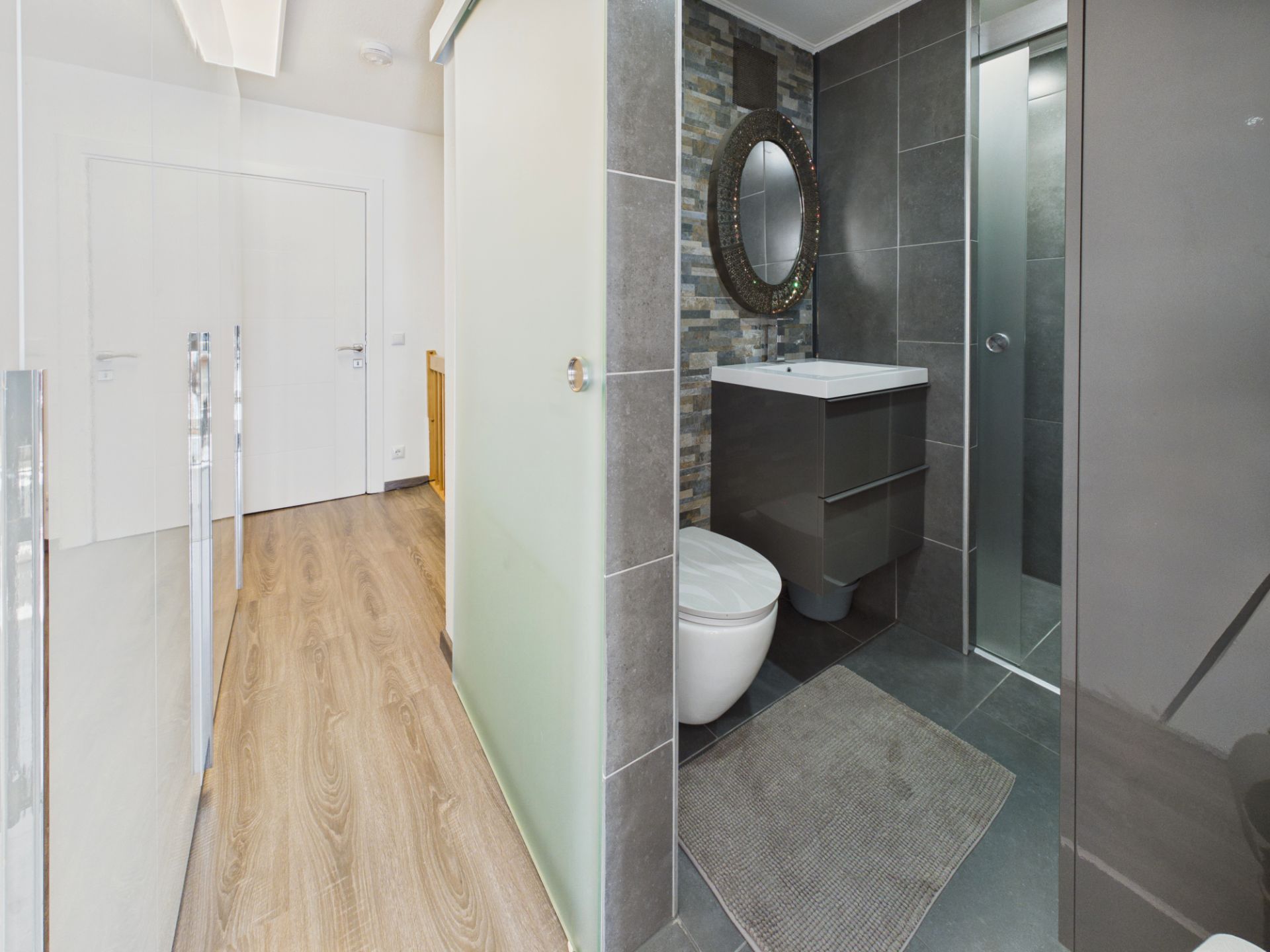 Modernes Badezimmer mit WC, Waschbecken, rundem Spiegel und Dusche, daneben Flur mit Holzboden.