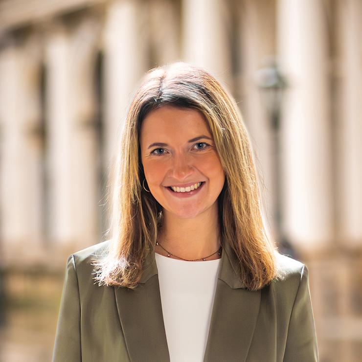 Frau mit mittellangen Haaren und grünem Blazer steht vor unscharfem Hintergrund.