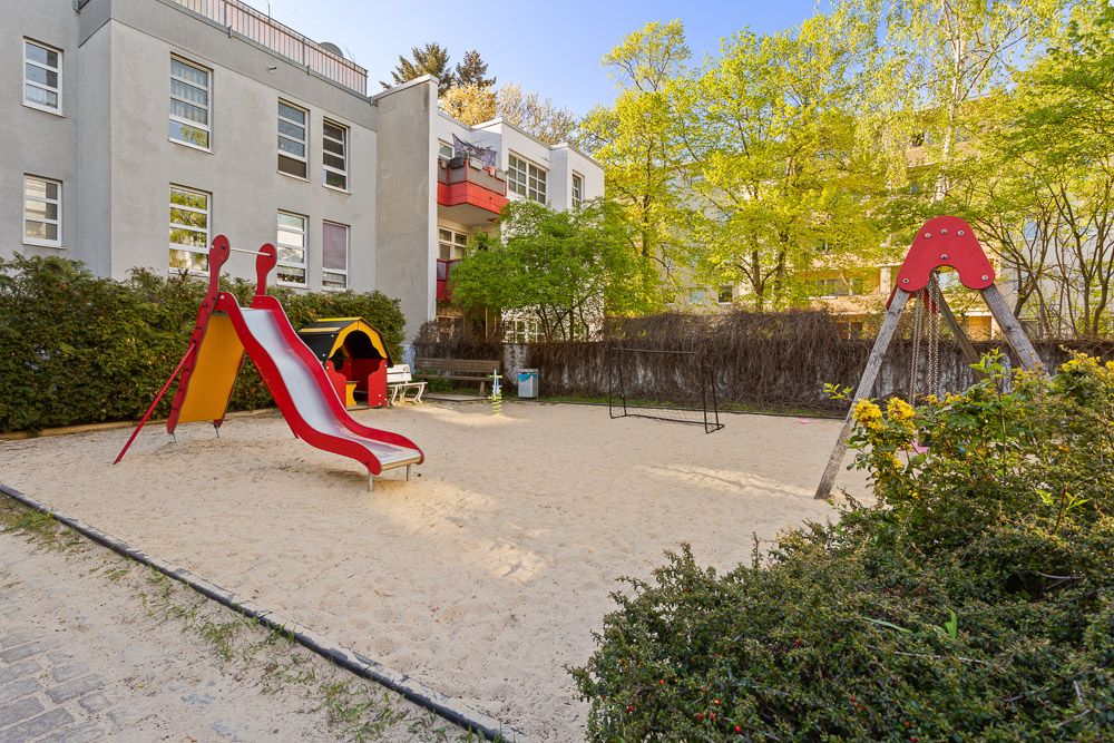 Spielplatz mit roter Rutsche, Schaukel und Spielhaus im Sand, umgeben von Wohngebäuden und Bäumen.