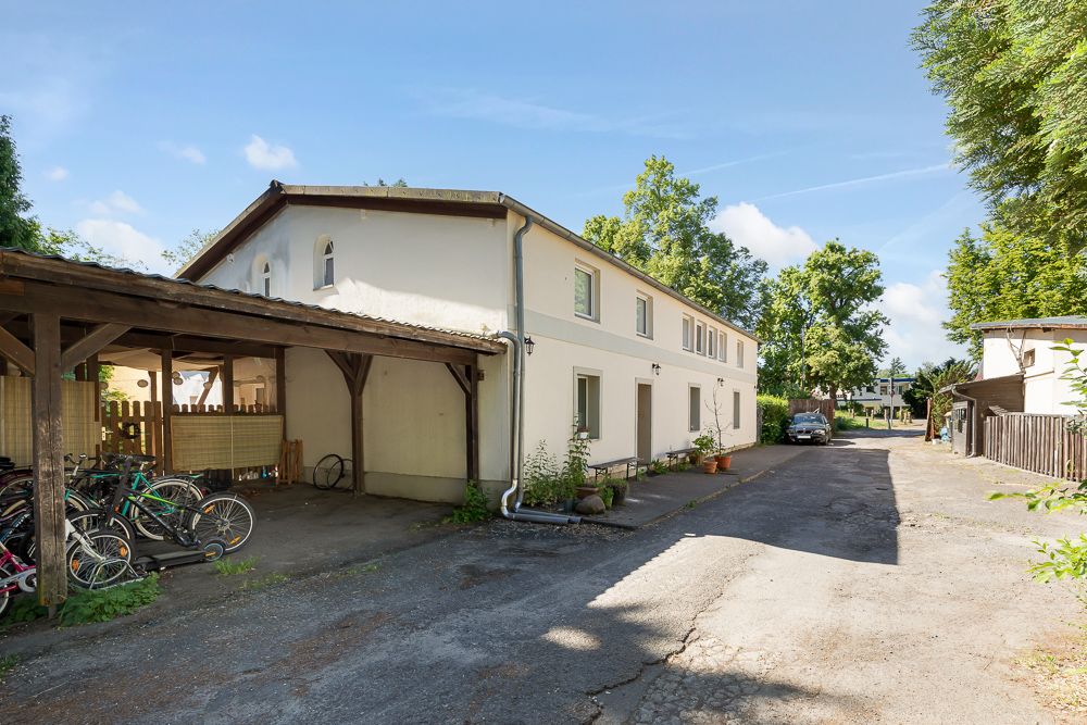 Zweistöckiges Wohnhaus mit Carport und mehreren Fahrrädern davor, daneben ein Hof mit Bäumen.