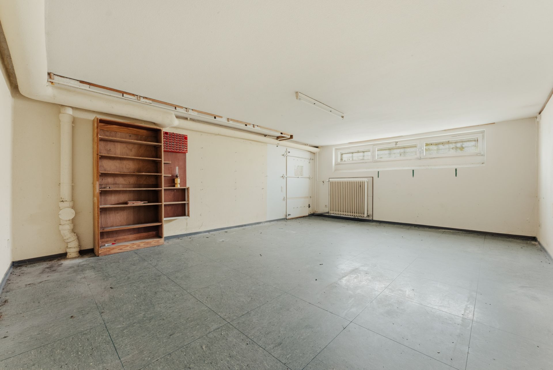 Leerer Kellerraum mit Regal, Fenster und Heizkörper an der Wand.