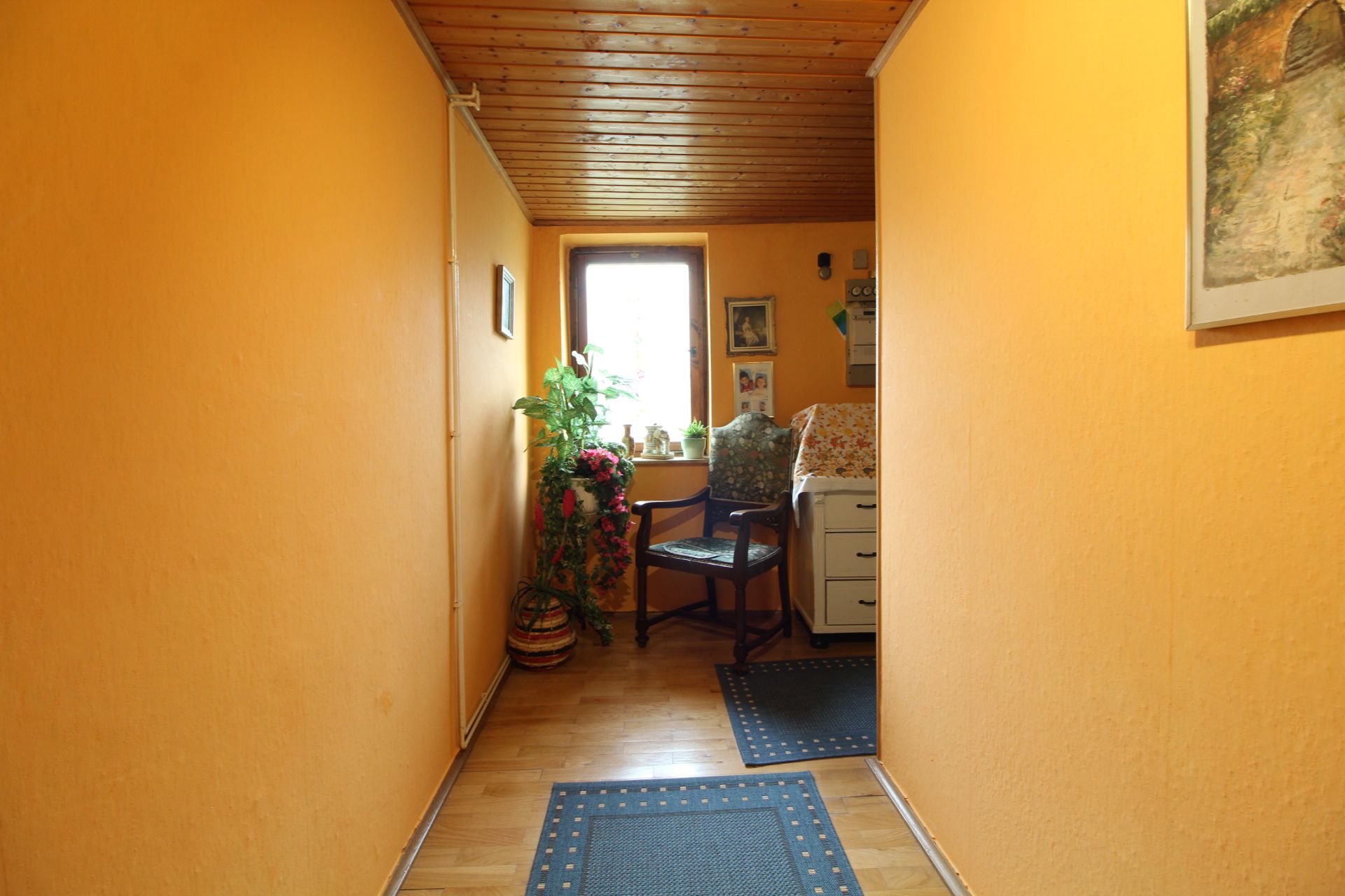 Flur mit gelben Wänden, Holzdecke, einem Stuhl, Pflanzen und einem Fenster am Ende.