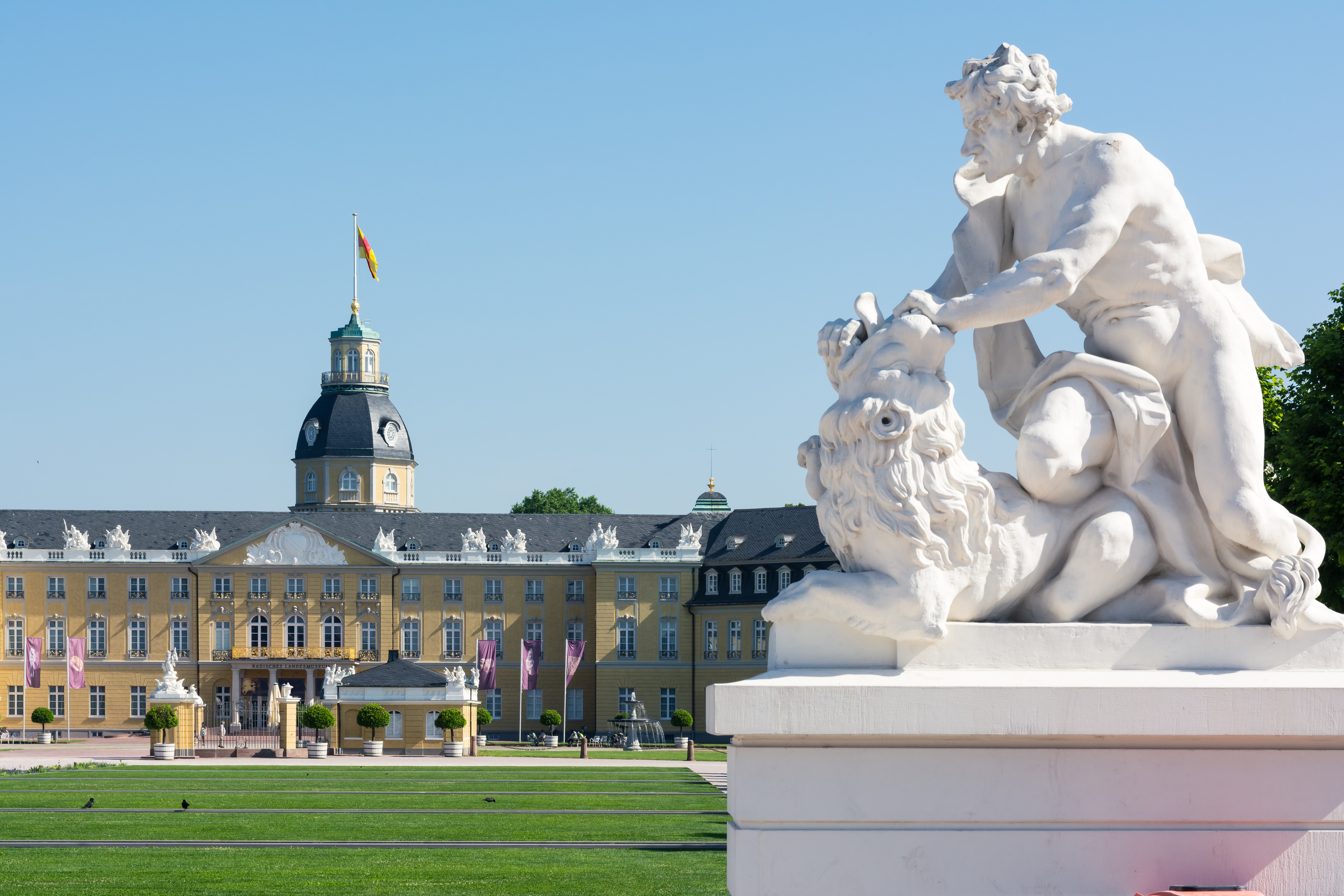 Großes gelbes Gebäude mit Turm und Statue eines Mannes mit Löwen im Vordergrund.