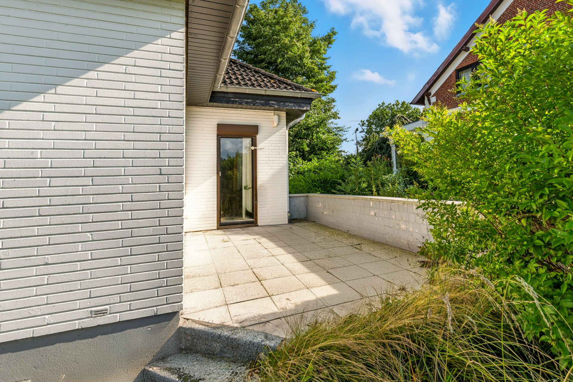 Gepflasterte Terrasse mit niedriger Mauer an einem weißen Haus, umgeben von Pflanzen.