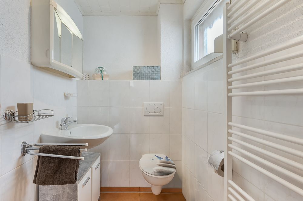 Kleines, helles Badezimmer mit Waschbecken, Wand-WC, Handtuchheizkörper und Fenster.