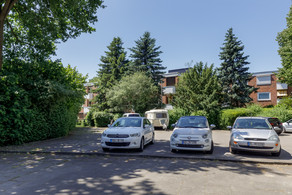 Drei Autos und ein Wohnwagen stehen auf einem Parkplatz vor einem Wohngebäude mit Bäumen im Hintergrund.