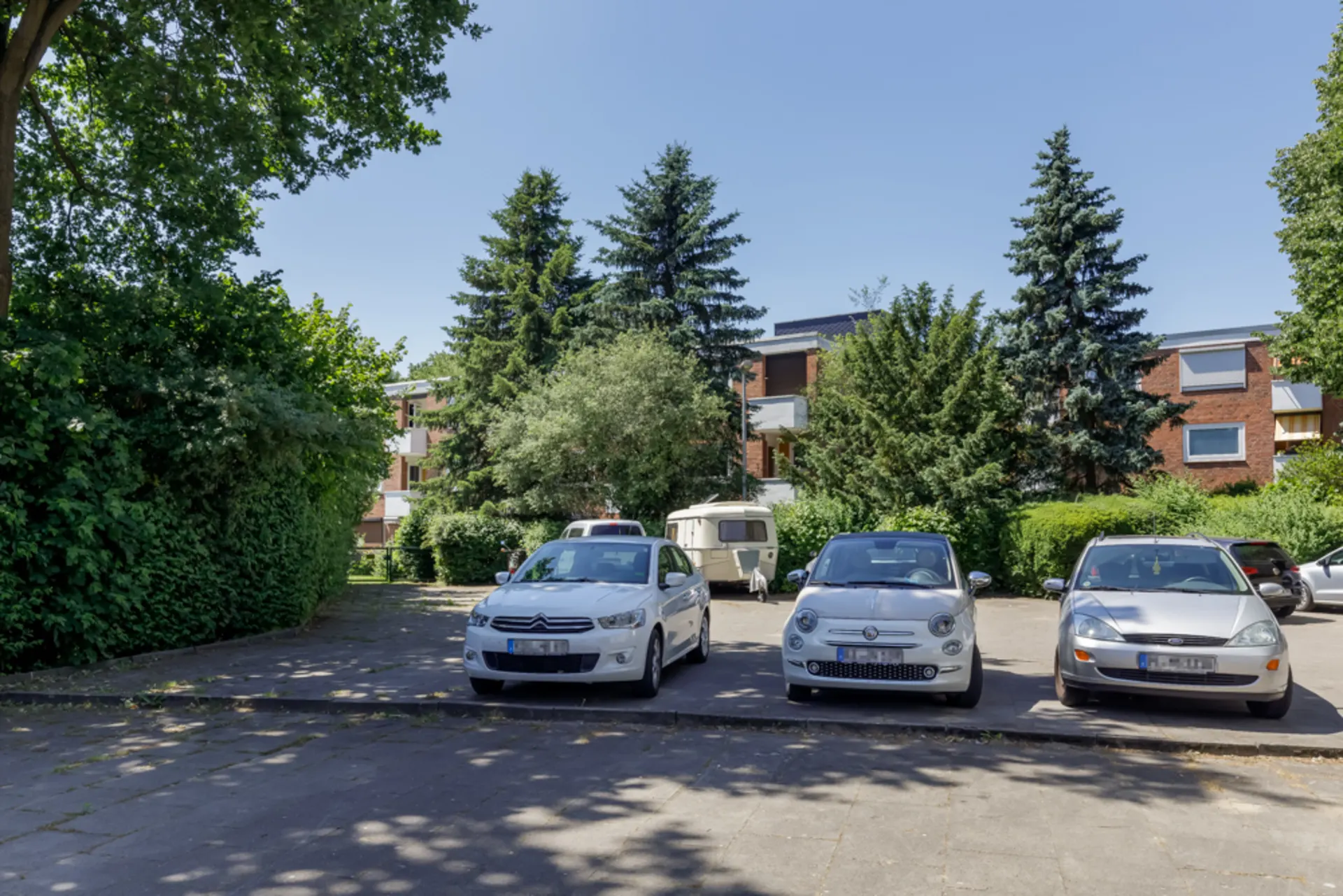 Drei Autos und ein Wohnwagen stehen auf einem Parkplatz vor einem Wohngebäude mit Bäumen im Hintergrund.