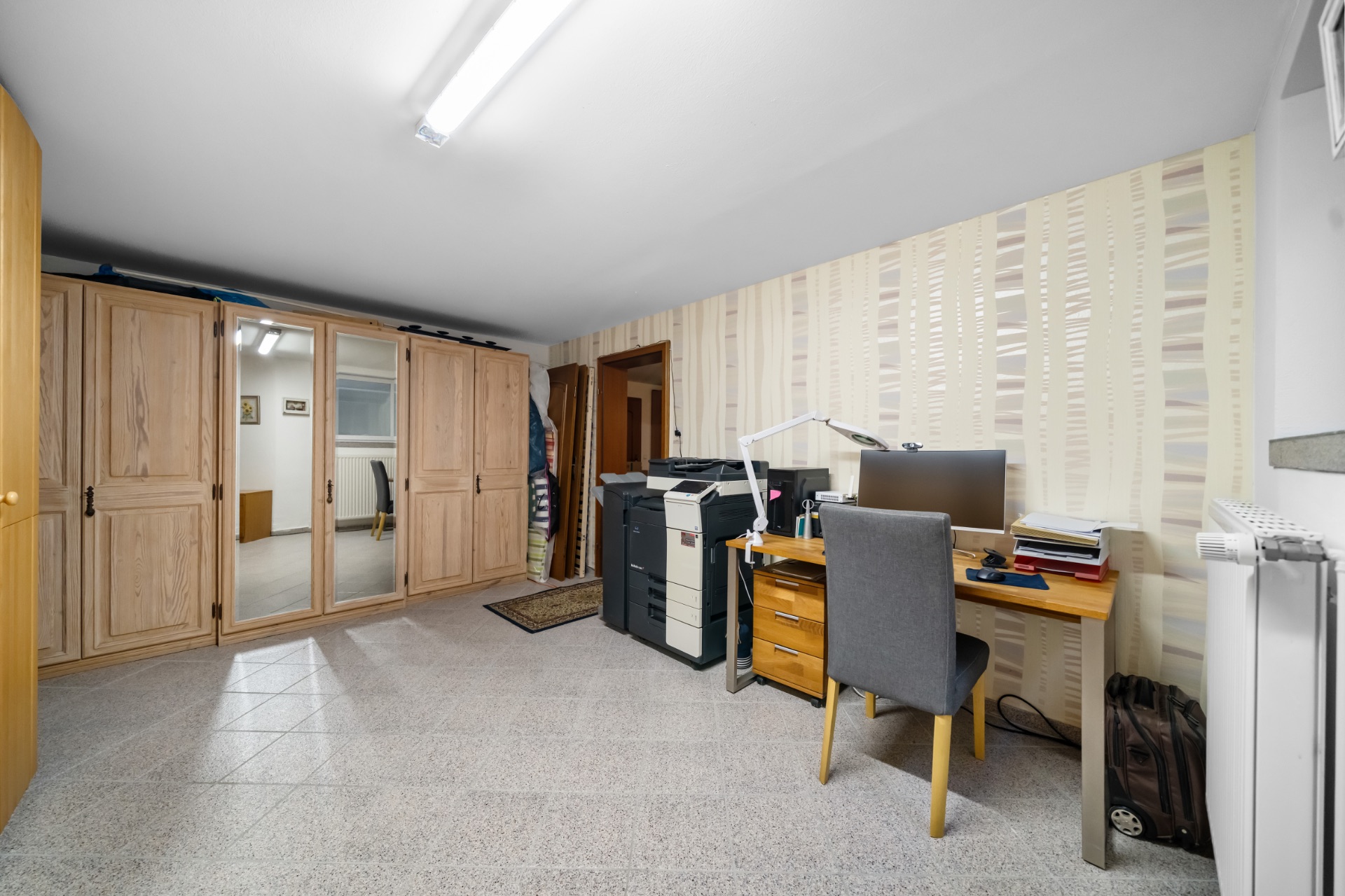 Arbeitszimmer mit Schreibtisch, Stuhl, Computer, Drucker und großem Holzkleiderschrank.