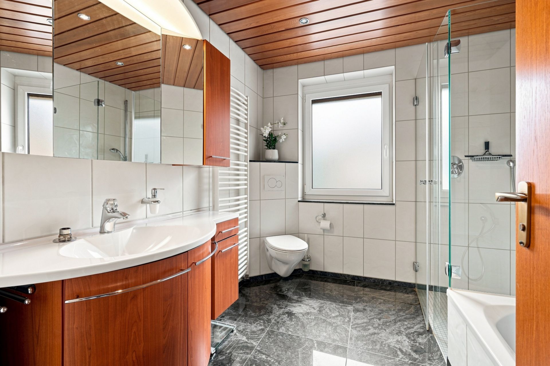 Modernes Badezimmer mit Holzdecke, großem Spiegel, Glasdusche, WC und Fenster.