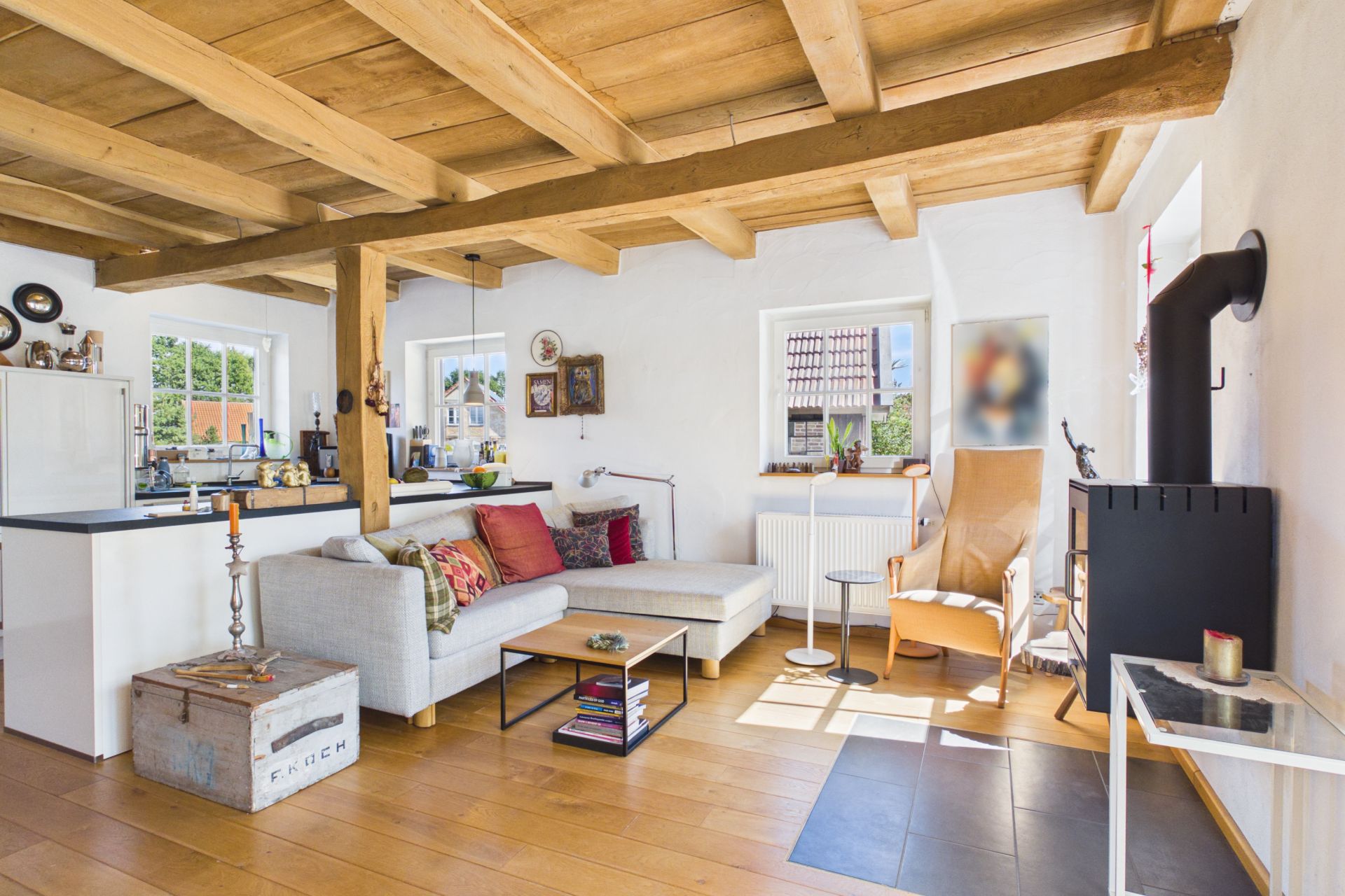Helles Wohnzimmer mit Holzdecke, Sofa, Sessel, Holzboden und Blick auf eine offene Küche.