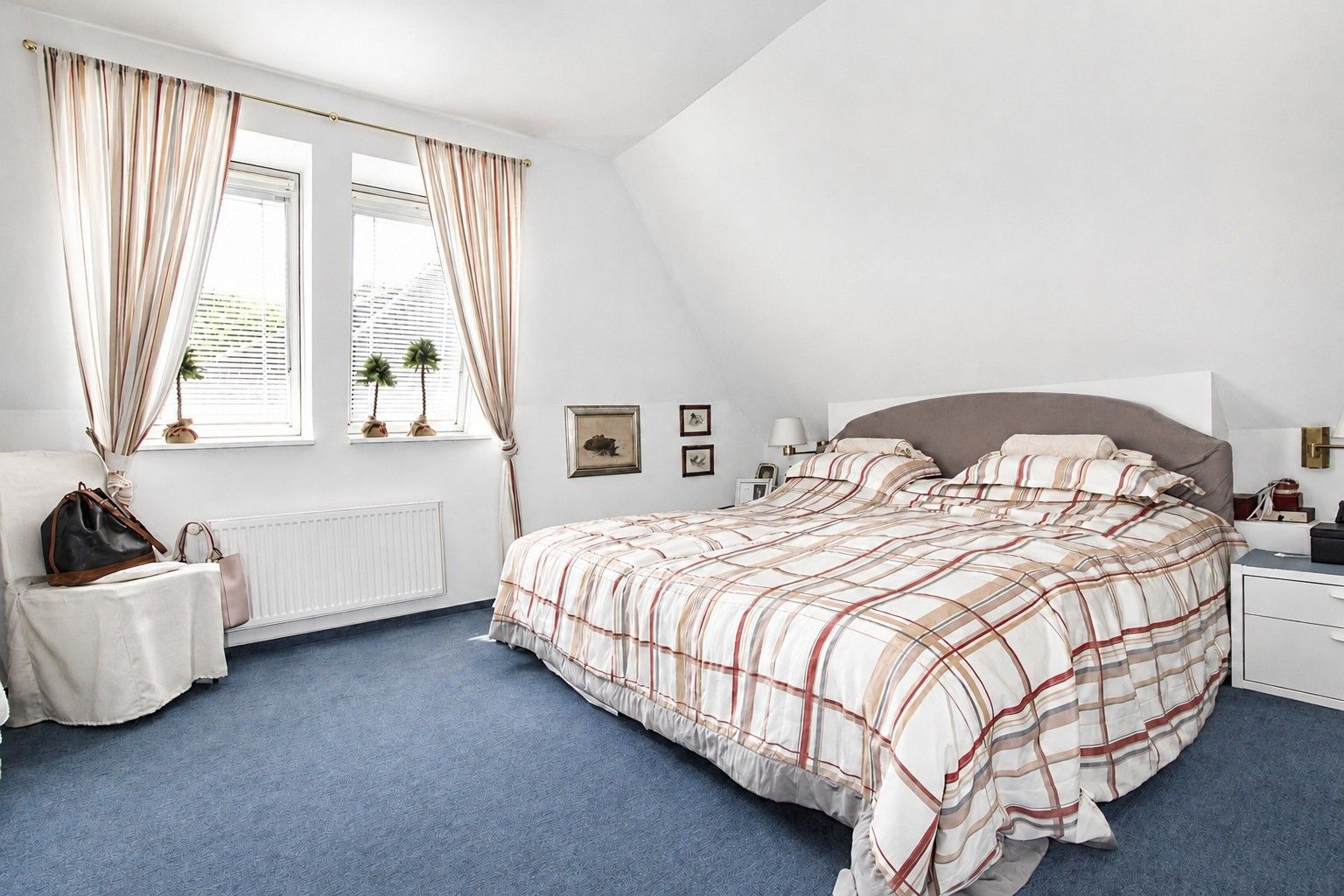 Helles Schlafzimmer mit Doppelbett, blauer Teppichboden und zwei Fenstern mit Vorhängen.
