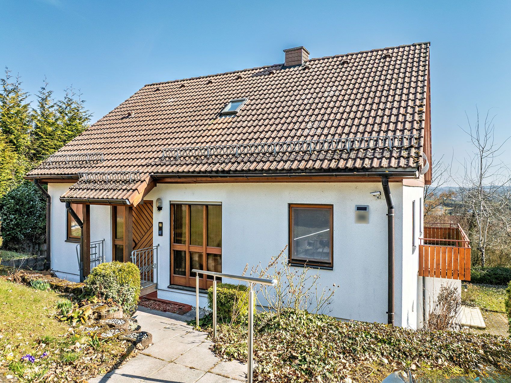 Einfamilienhaus mit Satteldach, weißer Fassade, Vorgarten und Holzveranda an einem sonnigen Tag.