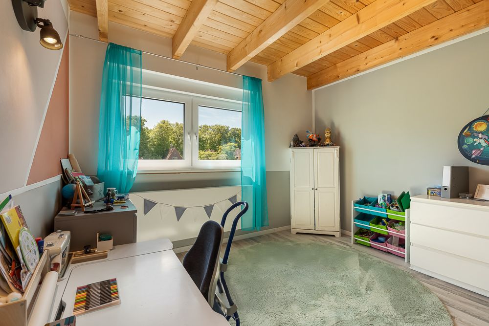 Helles Kinderzimmer mit Schreibtisch, Regal, Teppich und Holzbalkendecke.