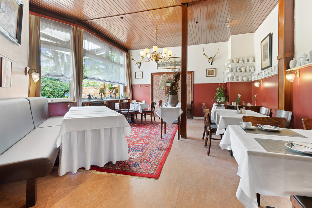 Heller Essbereich mit Holzmöbeln, Teppich, großen Fenstern und Tischen mit weißen Decken.