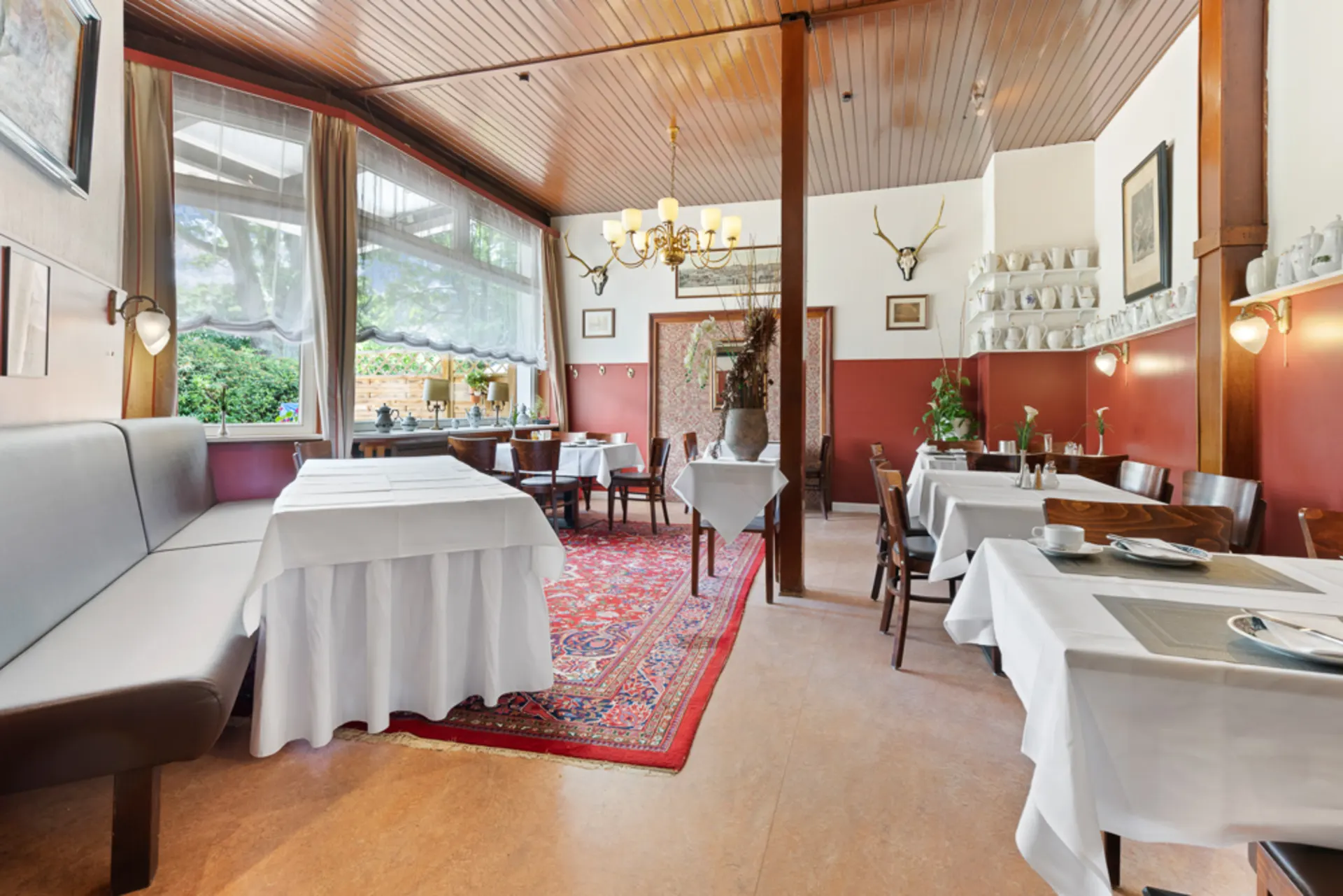 Heller Essbereich mit Holzmöbeln, Teppich, großen Fenstern und Tischen mit weißen Decken.